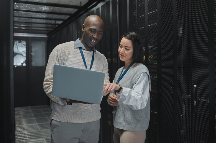 An IT support specialist holding a laptop, troubleshooting with a colleague in the server room